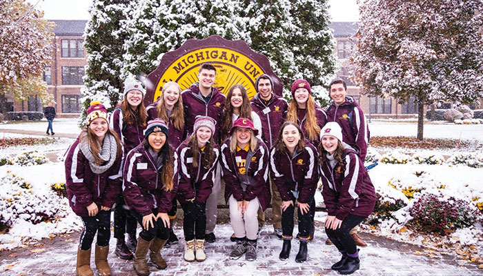A group of Central Michigan University ambassadors, wearing maroon and gold jackets, pose in the snow in front of the university seal.