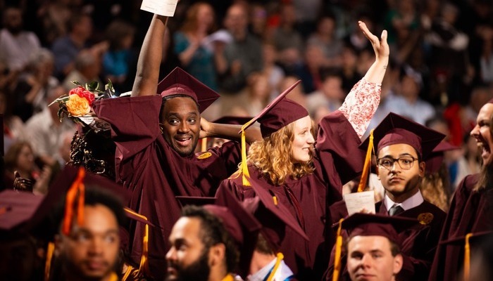 Diverse group of CMU graduates in maroon caps and gowns raising hands in celebration at a commencement ceremony.