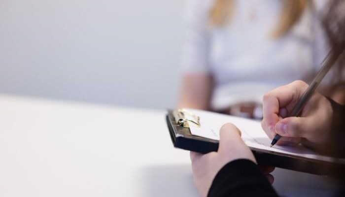 A person writing with a pen on a document attached to a clipboard, while another person sits in the background.