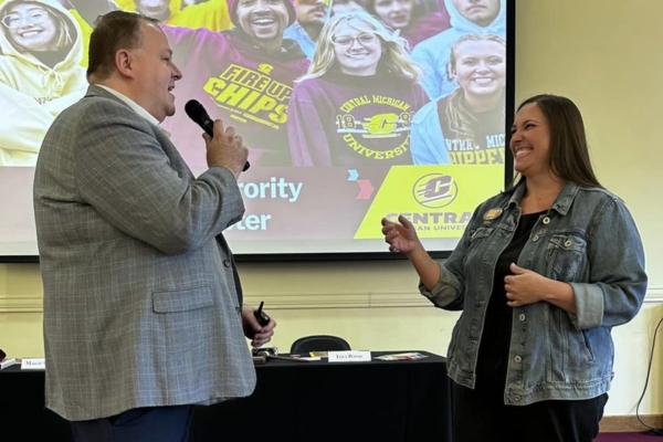 a man and woman talking into a microphone and standing in front of a projector screen.