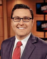 Greg Angel previous headshot for Alumni Awards wearing a purple suit jacket and red tie with white polka dots smiling with teeth with short brown hair and glasses.