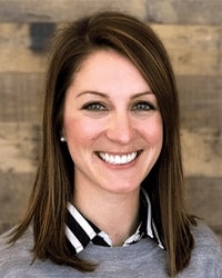 Lindsay Barnett's previous headshot for Alumni Awards wearing a gray sweater with a black and white collared shirt underneath and she has shoulder-length brown hair and is smiling with teeth.