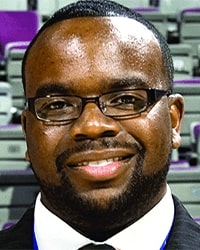 Lester Booker Jr.'s previous headshot for the Alumni Awards wearing a black suit with a white collar shirt black glasses and short black hair.