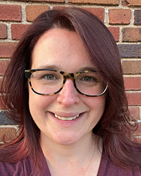 Caitlyn Demsky's Updated Headshot Photo- Dark red/purple died hair, side part, green/brown eyes black and brown glasses, smile with teeth, maroon v neck shirt, and brick background