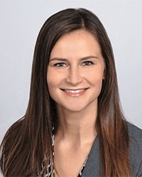 Kayla Davis's previous headshot for the Alumni Awards where she is smiling with teeth wearing a dark gray blazer and has long brown hair.