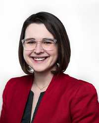 Caitlin Demsky's previous headshot for the Alumni Awards where she has short dark brown hair and clear glasses and is smiling with teeth while wearing a black top under a red jacket.