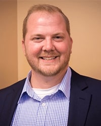 Dan Ekonen's headshot for the Alumni Awards where he is smiling with teeth with short blonde hair and is wearing a blue and white button down shirt and dark navy blazer.