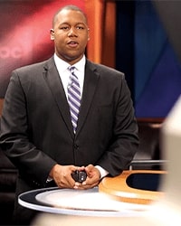 Matt Franklin's previous headshot for the Alumni Awards wear he is delivering a broadcast while wearing a black suit with a white and blue striped tie.