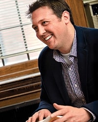 Dan Gaken's previous headshot for the Alumni Awards where he is looking off to the side and wearing a dark blue blazer and a white and blue plaid shirt and has short brown hair.