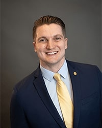 Mike Greene's updated headshot for the Alumni Awards where he is wearing a dark navy blazer with a light blue button-down shirt with a gold tie and has short brown hair and is smiling with teeth.