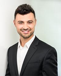 Derek Hirschman's headshot for the Alumni Awards with short black hair and smiling with teeth while wearing a black suit jacket and white button-down shirt.