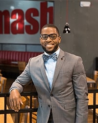 LaMarcus D. Howard's headshot for the Alumni Awards where he is wearing a gray suit with a light blue button-down underneath and a blue bowtie with black glasses and while smiling with teeth.