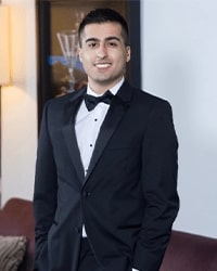 Nasir Hussain's headshot for the Alumni Awards with short black hair while he is wearing a black tuxedo and smiling with teeth.