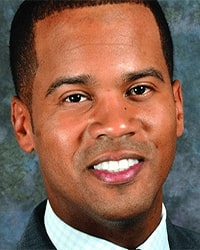 Lorron James's headshot for the Alumni Awards wearing a light blue and white plaid shirt with a black suit jacket and he is smiling with teeth.