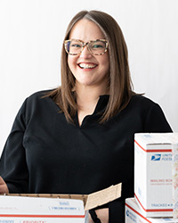 Jessi Ekonen's Updated Headshot Photo- Shoulder length brown hair, brown patterned round glasses, smile with teeth, red lipstick, long sleeve black dress top with v-neck, priority mail packages around individual, and white background