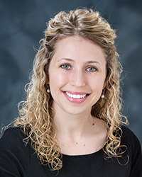 Kasee Stratton's Updated Headshot Photo- Blonde shoulder length curly hair, half hair is pulled back, green/blue eyes, light pink lipstick, smile with teeth, dangling pearl earrings, swoop neck black shirt, and gray background