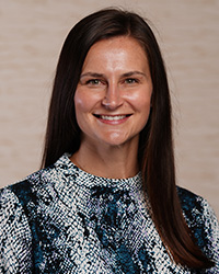 Kayla Davis' Updated Headshot Photo- Long dark brown hair, smile with teeth, red lipstick, high neckline patterned top, navy blue and white, and light tan background