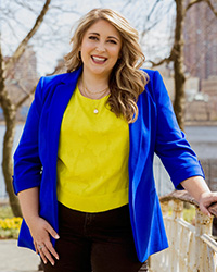 A woman standing photo, medium length blonde curling hair, red lipstick, smiling with teeth, yellow under shirt, electric blue blazer with sleeves rolled up partially, black dress pants, silver ring, layered necklace.