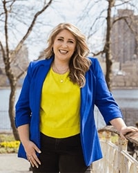 Kelley Kosuda's headshot for the Alumni Awards where she is wearing a bright blue blazer with a yellow blouse underneath and has long blonde hair while smiling with teeth standing in front of a river.