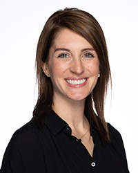 A woman with medium length brown hair, side hair part, green eyes, smile with teeth, silver stud earrings, simplistic necklace with a circular charm, black button up shirt with a collar, and a white background.