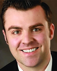 Michael Love's headshot for the Alumni Awards where he is wearing a black suit with a white collared shirt and has short brown hair and is smiling with teeth.