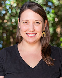 A woman with shoulder-length brown hair, dangling upside down U-shaped gold earrings, brown eyes, smile with teeth, pink lipstick, black v-neck short sleeve t shirt, and blurry green background.