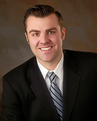 A man with short dark brown hair, green eyes, smile with teeth, white button up dress shirt with collar, black and light blue diagonally striped long tie, black suit coat, and brown/gray background.