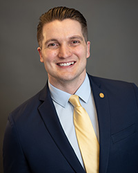 A man with short brown hair, brown eyes, long yellow tie, light blue button down shirt with a collar, navy blue suit coat, small circular yellow pin on the coat, and a grey background.