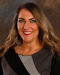 A woman with long dark brown hair, hoop earrings, smile with teeth, red lipstick, blue eyes, dark grey and black striped long sleeve shirt, black eyeliner and mascara, and brown photo backdrop background.