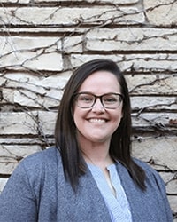 Jesi Parker-Ekonen's previous headshot for the Alumni Awards where he is standing in front of a white brick wall wearing a blue cardigan and light blue blouse and has long brown hair and black glasses while smiling with teeth.