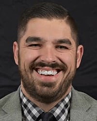 Tyler Parson's previous headshot for the Alumni Awards where he is wearing a black and white plaid shirt with a gray suit jacket and black tie and has short black hair and is smiling with teeth.