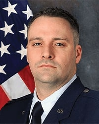 Steve Poe's previous headshot for the Alumni Awards where is wearing an Air Force uniform in front of an American flag and has short black hair with a neutral expression.