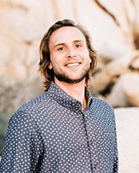 Zachary Roszczewski's headshot for the Alumni Awards where he is standing outside wearing a navy shirt with white polka dots and he is smiling with teeth with longer blonde hair.