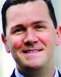 Mike Schuette's headshot for the Alumni Awards and he is wearing a dark suit with a light blue collared shirt and he has short brown hair and is smiling with teeth.