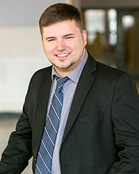 A man with dark brown hair, a smile with teeth, goatee, blue button down dress shirt with collar, long navy blue tie with white diagonal stripes, black dress coat, and hallway background.