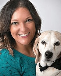 Natalie Shattuck's previous headshot for the Alumni Awards where she is with her dog and is wearing a dark teal top with black polka dots and has shoulder-length brown hair and is smiling with teeth.