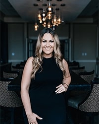 Courtney Stanley in a black dress standing in front of a chandelier with long blonde curled hair.