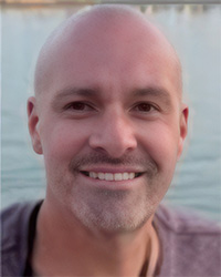 Steve Poe Updated Headshot- gray beard, smile teeth, maroon short sleeve shirt, dark brown eyes, light blue water background