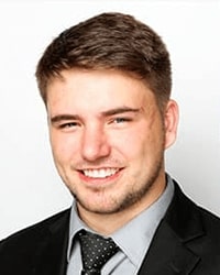 Scott Stewart's previous headshot for the Alumni Awards where he is wearing a black suit with a gray button-down shirt and a black tie that has white polka dots and he is smiling with teeth and has short brown hair.