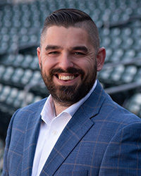 Tyler Parsons dark brown hair, dark brown beard, smile with teeth, white button up shirt with color, navy blue dress coat with light blue stripes, dark green bleacher seating background.