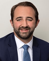 Drew Weil's headshot for the Alumni Awards where he is wearing a dark navy suit with a white button-down shirt and a blue and white polka dot tie and he has short brown hair and is smiling with teeth.