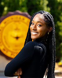 Headshot of 2024 Alumni Awards Future Alumni Leader Recipient Aaliyah Howard standing in front of the CMU seal and she is wearing a long-sleeve black shirt and has long black hair in braids and is smiling with teeth.