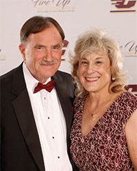 Headshot of 2024 Alumni Award Dick Enberg CMU Alumni Commitment Recipients Mike and Pam Murray and Mike is standing left in black tux and maroon bow tie and Pam is standing right wearing a maroon and white dress and they are smiling with teeth.