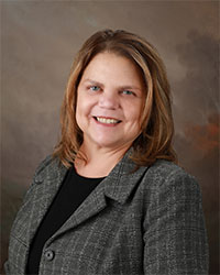 Headshot of 2024 Alumni Award Distinguished Alumni Recipient Melinda Coffin who is wearing a dark gray pinstriped blazer with a black shirt and she has shoulder-length light brown hair and is smiling with teeth.