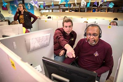 Two people in a cubicle are wearing maroon and looking at a computer.