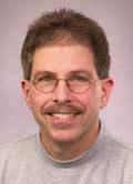 This is a headshot of a man wearing a gray mock neck shirt with light brown hair a mustache and glasses.