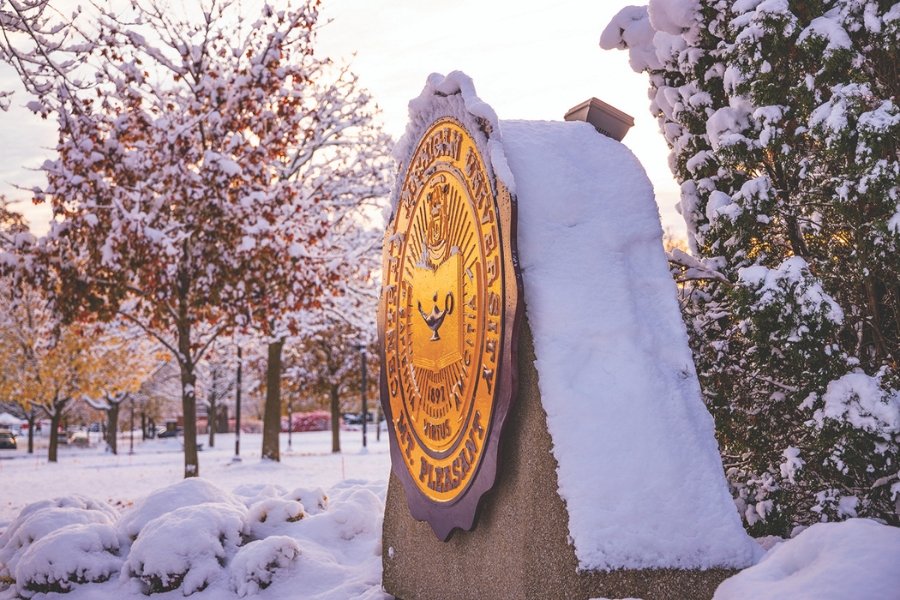 A dusting of white snow covers the maroon and gold CMU Seal and surrounding nature outdoors.