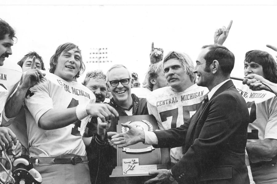 This is a black and white photo of the CMU football team receiving a trophy. A man dressed in a suit is offering his hand to one of the players for a handshake while others gather in celebration in the background.