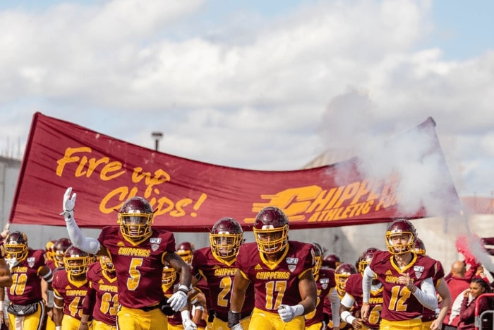 CMU Football team running onto the field. There is smoke and a large maroon and gold banner.