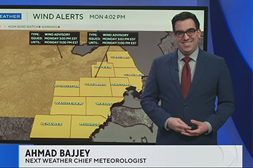 A man with dark hair wearing a suit and tie stands in front of a weather map on a TV screen that says wind alerts.